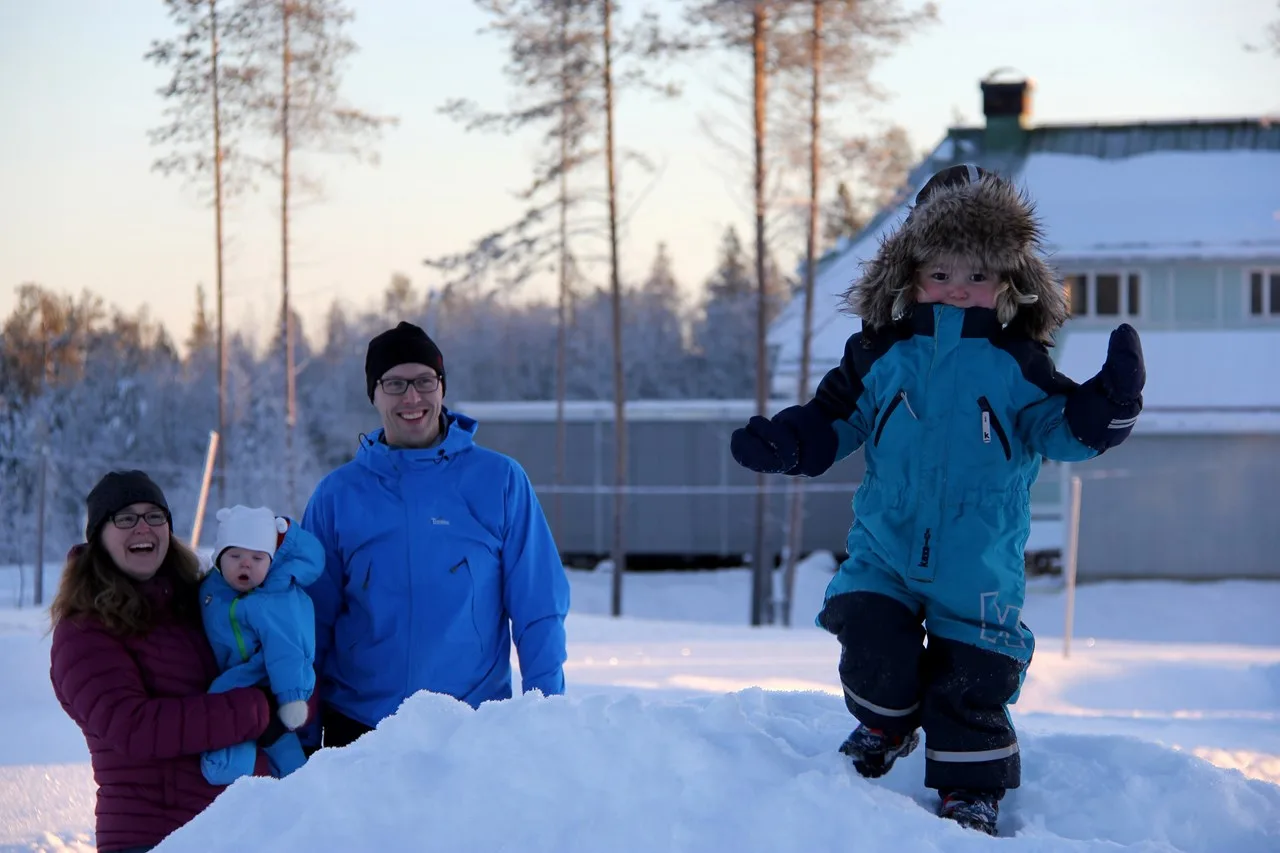Barn klädd i overall står på en snöhög omringad av sin familj.